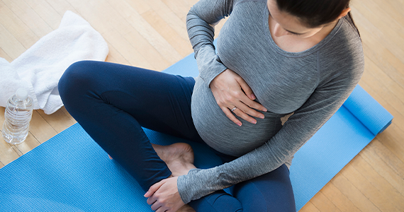 A pregnant woman in athletic clothes sits cross legged on a yoga mat. Her hand rests on her belly as she takes a break