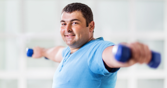 A middle-aged man smiles as he exercises. He holds weights away from his body and has broken a light sweat.