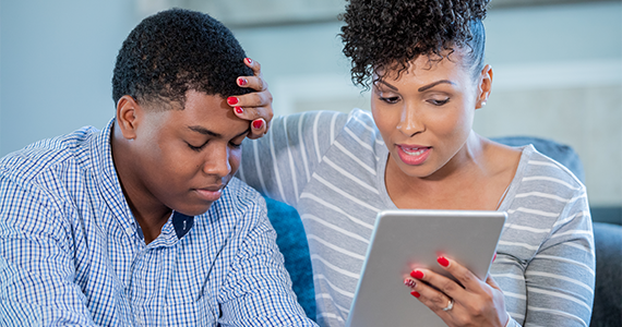 A patient and caregiver with a hand on the patient's forehead participate in a medical consultation using a tablet.