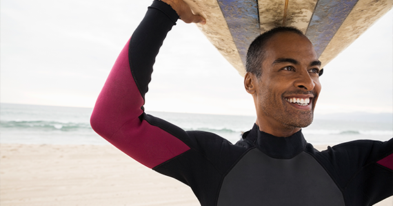 Un hombre sonriente está usando su tiempo libre remunerado para correr algunas olas.  Lleva traje de neopreno y sostiene su tabla de surf sobre la cabeza.